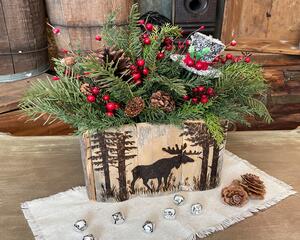 This shows a handmade wooded box that has a moose and pine trees wood-burned on the front. The box hold a Christmas greenery arrangment. 